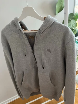 Grå zip hoodie från Burberry XL - Snygg grå hoodie från Burberry i storlek XL. Dragkedja framtill, två stora fickor och klassisk huva med dragsko. Liten Burberry-logga broderad på bröstet. Tillverkad i mjukt bomullsmaterial som känns riktigt skönt mot huden.