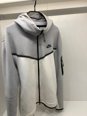 Nike tech fleece  - Nike hoodie med dragkedja i grått och vitt. Stora färgblock, svart Nike-logga på bröstet och svarta detaljer längs dragkedjan och ärmslut. Huva och ficka med dragkedja på ena ärmen. Tillverkad i mjukt sweatshirtmaterial, perfekt för chill eller träning.