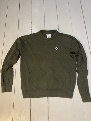 Olivgrön crewneck från Junkyard - Säljer en olivgrön crewneck från Junkyard med rund halsringning och ribbade muddar. Tröjan har en liten broderad logga på bröstet och är tillverkad i mjukt material. Perfekt för dig som gillar enkel och stilren streetwear.