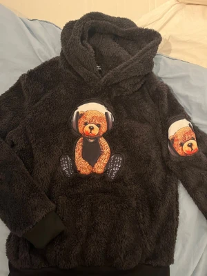 Mjuk svart hoodie med nalletryck - Säljer en fluffig svart hoodie med stor huva och tryck av en nallebjörn med hörlurar på bröstet och ärmen. Hoodien har muddar vid ärmslut och nederkant samt en stor magficka. Perfekt för dig som gillar mysiga och lekfulla plagg.