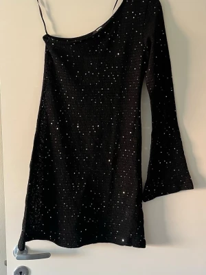 Svart glittrig klänning med paljetter - Säljer en svart kortklänning med glittriga paljetter och en axelbandslös design på ena sidan. Klänningen har lång ärm på ena armen och är tillverkad i ett stretchigt material som ger en snygg passform. Perfekt för dig som vill sticka ut med en unik och trendig look.