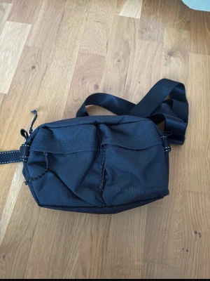 Svart axelväska från Peak Performance - Snygg svart axelväska från Peak Performance med två framfickor och justerbar axelrem. Väskan är gjord i slitstarkt tyg och har detaljer i svart med vita sömmar. Perfekt för dig som vill ha en praktisk och stilren väska till vardagen.