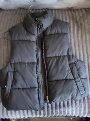 Svart dunväst från 157 - Snygg svart dunväst från 157 med hög krage och dragkedja framtill. Västen har två sidofickor och quiltad design som ger en modern look. Perfekt att slänga över en hoodie eller tröja för extra värme. Materialet är mjukt och lätt, vilket gör den enkel att bära.