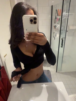 Svart topp med knyt och v-ringning - Säljer en svart omlottopp med lång ärm och djup v-ringning. Toppen har en croppad passform och knyt i sidan som ger en snygg siluett. Perfekt att styla med höga jeans eller kjol för en trendig look.🥰