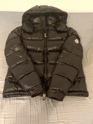 Svart dunjacka från Moncler med huva - Säljer en svart dunjacka från Moncler med glansig finish och klassisk Moncler-logga på ärmen. Jackan har huva, dragkedja framtill och breda, vadderade paneler som ger en riktigt puffig look. Perfekt för kalla vinterdagar.