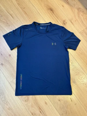 Under Armour träningströja - Tröjan har en liten fläck där man kan se i bild 3                                                            Snygg blå funktions-tshirt från Under Armour med korta ärmar och rund hals. Tröjan har diskret logga på bröstet och textdetaljer på ärmen och nederkanten. Tillverkad i ett lätt och snabbtorkande material som passar perfekt till träning.