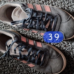 Adidas sneakers grå/rosa strl 39 - Snygga Adidas sneakers i grå mocka med klassiska rosa ränder på sidorna och mörkgrå snören. Låg modell med vit sula och retrovibe. Perfekta för dig som gillar streetstyle och vill ha något bekvämt och stilrent.