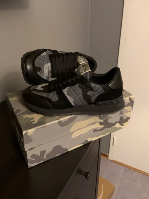Valentino svart/grå camo sneakers - Säljer ett par snygga Valentino sneakers med svart och grått camo-mönster. Skorna har svarta detaljer i mocka och mesh, samt robust svart sula. Kommer med originalkartong, extra skosnören och dustbag. Perfekta för dig som gillar streetwear och exklusiva märken.