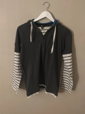 Svart hoodie med ränder från Elanino - Cool svart hoodie från Elanino med vita och svarta ränder på ärmarna och huva med dragsko. Tröjan har knappar vid halsen och kontrastfärgad blå insida i huvan. Perfekt för en avslappnad och trendig stil.