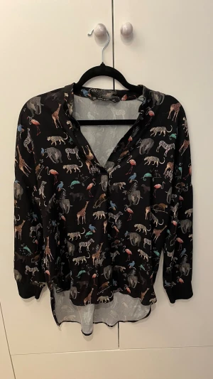 Svart blus med djurmönster - Snygg svart blus med färgglatt djurmönster som visar elefanter, zebror, giraffer och flamingos. Blusen har långa ärmar med muddar och en avslappnad passform. Perfekt för dig som gillar unika prints och vill sticka ut lite extra.