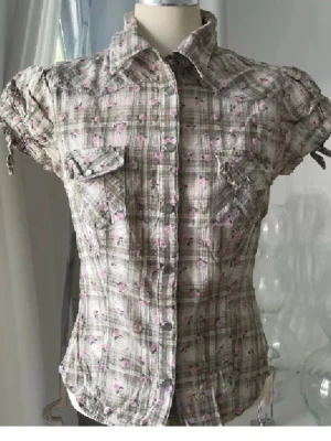 Rutig kortärmad skjorta med blommor - Kortärmad skjorta i beige och grönt med rutigt mönster och små rosa blommor. Skjortan har knappar framtill, två bröstfickor med lock och knappar samt snörning på ärmarna. Perfekt för dig som gillar en söt och avslappnad stil.