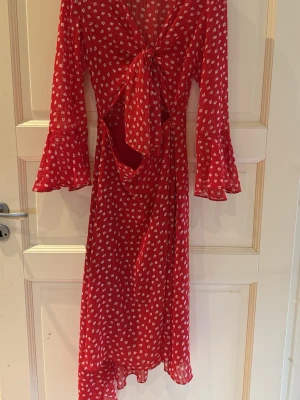 Röd blommig långklänning från Adoore - Säljer en röd långklänning från Adoore med vita blommor, v-ringning, knyt framtill och bar i midjan. Klänningen har långa ärmar med volang vid ärmslut och är tillverkad i ett lätt, luftigt material. Perfekt för sommarens alla tillfällen!