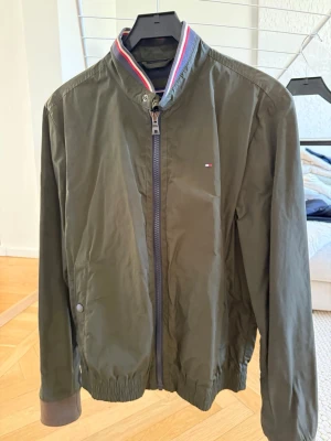 Olivgrön bomberjacka Tommy Hilfiger - Snygg olivgrön bomberjacka från Tommy Hilfiger med dragkedja framtill och ribbade muddar. Jackan har en klassisk ståkrage med röd, vit och blå rand samt liten logga på bröstet. Perfekt för dig som vill ha en stilren och tidlös look.