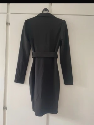 Svart kavajklänning med bälte - Stilren svart kavajklänning från newyorker med lång ärm och markerad midja. Klänningen har klassisk krage och omlott-look framtill, vilket ger en elegant och modern känsla.