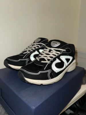 Svarta B3O sneakers med vit logga - Säljer ett par svarta sneakers med vit och reflekterande logga på sidan. Skorna har vita snören och en vit, chunky sula med detaljer. Finns dock lite skador på yttersulan och fläck på sulans kant. Materialet är mesh och syntet, vilket ger en sportig och modern look.