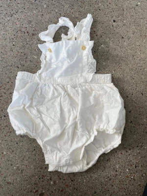 Vit babyromper med volanger H&M - Söt vit babyromper från H&M i ekologisk bomull. Rompern har volangdetaljer på axelbanden, små knappar framtill och resår vid benen för en bekväm passform. Perfekt för en gullig och stilren look till de minsta.