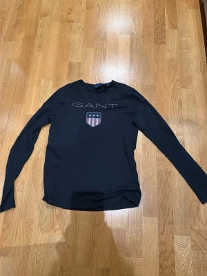 Svart långärmad topp från Gant - Snygg svart långärmad topp från Gant med tryckt logga och sköld på bröstet. Klassisk rund halsringning och mjukt material som känns skönt mot huden. Perfekt till jeans eller kjol för en clean och stilren look.