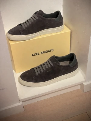 Axel Arigato grå mocka sneakers - Snygga sneakers från Axel Arigato i mörkgrå mocka med matchande grå skosnören och vit sula. Minimalistisk design med  logga i guld på sidan. Lågt skaft och klassisk siluett som passar till det mesta. Perfekta för dig som gillar stilrena och trendiga skor. Skorna är endast använda 2 gånger för att de var för små. Skorna är endast lite smutsiga på undersidan. Priset går såklart att diskutera och ställ gärna frågor! 
