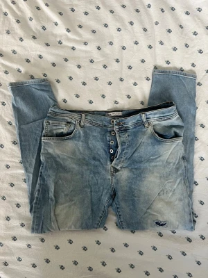 Ljusblå jeans med knappar från ZARA - Säljer ett par ljusblå jeans från ZARA med knäppning framtill och klassiska fem fickor. Jeansen har en snygg tvättad look och är raka i modellen. Perfekt för dig som gillar en avslappnad och trendig stil. Ungefärliga mått (platta): Längs midjan: 45 cm Innerbenslängd: 69cm  Ytterbenslängd: 101 cm