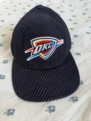 OKC keps från New Era - Snygg svart Oklahoma City Thunder NBA lag keps med OKC-logga broderad framtill och orangea detaljer på skärmen. Märkt med New Era-logga på sidan. Klassisk form och rak skärm, perfekt för dig som gillar basket och streetwear.