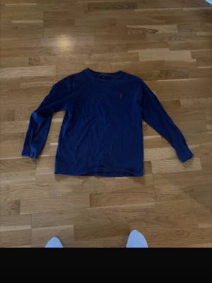 Mörkblå långärmad tröja Polo Ralph Lauren - Mörkblå långärmad tröja från Polo Ralph Lauren med klassisk röd broderad logga på bröstet. Tröjan har rund halsringning och är tillverkad i mjukt bomullsmaterial. Enkel och stilren design som passar till det mesta.
