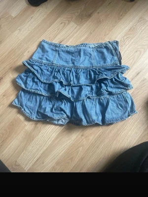 Blå volangkjol i jeans från Kappahl - Säljer en blå jeanskjol från Kappahl med flera lager volanger. Kjolen har en kort modell och är gjord i ett mjukt denimtyg som ger en avslappnad men ändå trendig look. Perfekt för dig som gillar en lekfull och cool stil.