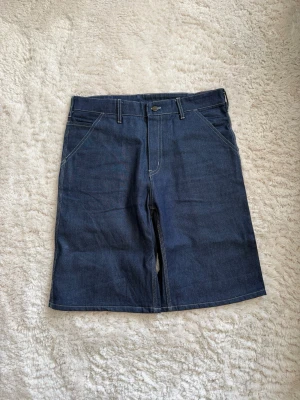 Mörkblå denimshorts från Carhartt - Säljer ett par mörkblå denimshorts från Carhartt med klassisk femficksdesign, bälteshällor och kontrastsömmar. Shortsen har raka ben och praktiska bakfickor, inklusive Carhartt-logga på ena fickan. Perfekta för en avslappnad streetwear-look. Köpte dem på en autentisk marknad i Bangkok, storlek står xl men det är barnstorlek, så om du är runt M så passar dem