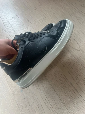 Svarta Philipp Plein sneakers - Stilrena svarta sneakers från Philipp Plein med vita sulor och detaljer i metallic. Ovandelen är i en mix av läder och mocka, med perforerade sidor och logga bak på hälen. Insidan är beige och skorna har klassisk snörning. Passar till allt möjligt även kostym! 