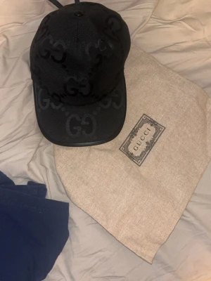 Svart keps från Gucci med GG-mönster - Säljer en svart keps från Gucci med det klassiska GG-mönstret i svart ton-i-ton över hela kepsen. Kepsen har böjd skärm och justerbar rem bak. Materialet är mesh-liknande och kepsen kommer med original dustbag.