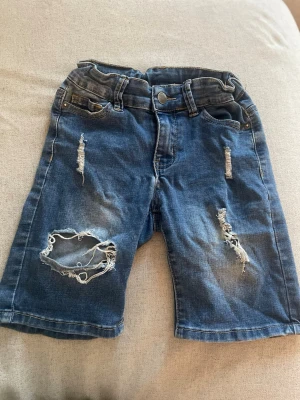 Blå slitna jeansshorts med hål Barn st 128 - Säljer ett par blå jeansshorts från H&M med slitna detaljer och hål på framsidan. Klassisk femficksmodell med knapp och dragkedja. Perfekta för en avslappnad och trendig look. Materialet är bomull med en skön denimkänsla. Barn med storlek 128