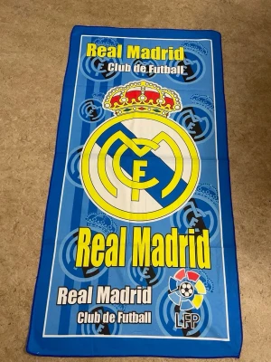 Real Madrid blå halsduk med logga - Snygg halsduk i blått med Real Madrids klubbmärke och texten 'Real Madrid Club de Futbol' i gult och vitt. Stor logga i mitten och LFP-märke längst ner. Perfekt för alla fotbollsfans som vill visa sitt stöd för laget.