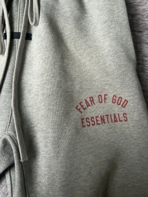 Grå mjukisbyxor Fear of God Essentials - Snygga grå mjukisbyxor från Fear of God Essentials med tryck i rött på ena benet. Byxorna har dragsko i midjan, resår vid benslut och är gjorda i mjukt bomullsmaterial. Perfekta för en chill och avslappnad stil.