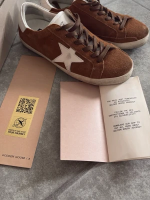 Bruna Golden Goose sneakers med stjärna - Snygga bruna sneakers från Golden Goose med vit stjärndetalj på sidan och vita hälpartier. Skorna har brunt mockaläder, bruna skosnören och en klassisk låg siluett. Perfekta för dig som gillar streetstyle och vill sticka ut med en ikonisk design. Finns en liten skada därav priset, går inte ner en enda krona. Går och fixa skadan med lim, annars är de helt nya och inte smutsiga och inga andra defekter alls