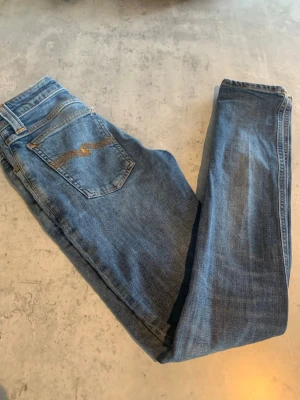 Blå jeans från Nudie Jeans - Snygga blå jeans från Nudie Jeans med klassisk femficksdesign och guldfärgad söm på bakfickan. Jeansen har rak passform och är tillverkade i denim av bomull. Perfekta för en avslappnad och trendig look.