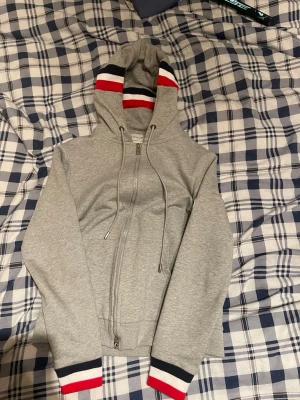 Grå Moncler hoodie med dragkedja - Snygg grå hoodie från Moncler med hel dragkedja och huva. Ränder i rött, vitt och marinblått på huva och ärmslut ger en sportig vibe. Klassisk Moncler-logga på ena ärmen. Perfekt för en chill och stilren look.