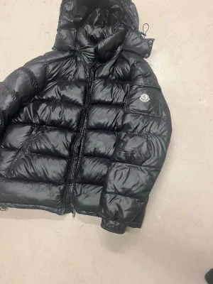 Moncler jacka - Snygg svart dunjacka från Moncler med glansig finish och klassisk pufferdesign. Jackan har huva, dragkedja framtill, två sidofickor med dragkedja och Moncler-logga på ärmen. Perfekt för kalla vinterdagar och riktigt trendig stil.