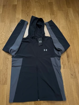 Svart och grå jacka  från Under Armour - Svart och grå jacka från Under Armour med hel dragkedja och justerbar huva. Tillverkad i ett lätt och funktionellt material, perfekt för träning eller chill. Under Armour-logga på bröstet ger en sportig vibe.
