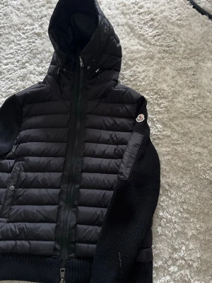 Svart pufferjacka från Moncler - Snygg svart pufferjacka från Moncler med quiltad front och rygg, stickade ärmar och huva med dragsko. Jackan har två fickor med tryckknappar och Moncler-logga på ena ärmen. Perfekt för kalla dagar och riktigt trendig stil.