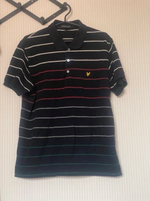 Randig pikétröja från Lyle & Scott - Snygg svart pikétröja från Lyle & Scott med tunna horisontella ränder i vitt, rött och grönt. Tröjan har korta ärmar, klassisk krage och knäppning framtill. Ikonisk gul logga broderad på bröstet. Perfekt för en avslappnad och stilren look.