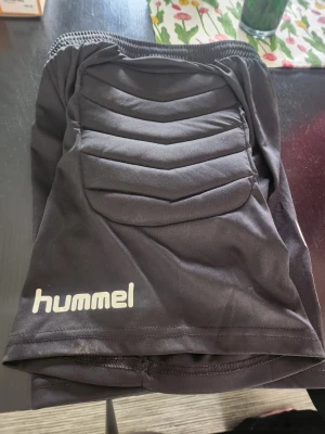 Svarta målvaktsshorts från Hummel - Svarta målvaktsshorts från Hummel med vadderade skyddspaneler på sidorna. Shortsen har elastisk midja och är tillverkade i ett slitstarkt, stretchigt material som passar perfekt för fotbollsträning eller match.