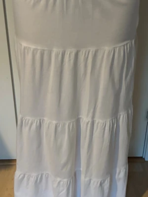 Vit långkjol med volangdetaljer - Säljer en vit långkjol med flera volangpartier som ger ett flowy och avslappnat intryck. Kjolen har en enkel och stilren design, perfekt för sommarens alla tillfällen. Tillverkad i mjukt bomullstyg för extra komfort.