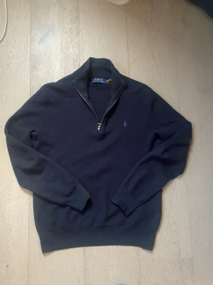Mörkblå halvzip-tröja från Polo Ralph Lauren - Mörkblå stickad tröja från Polo Ralph Lauren med halv dragkedja och broderad logga på bröstet. Tröjan har ribbade muddar vid ärmslut och nederkant samt hög krage. Perfekt för en stilren och klassisk look.