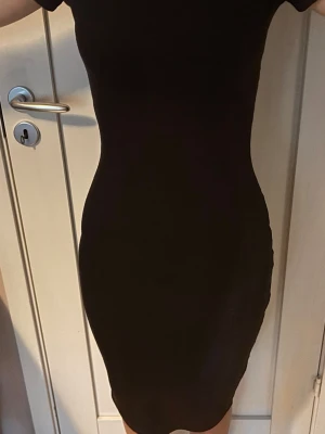 Svart tight kortklänning - En svart, tight kortklänning med korta ärmar och enkel, stilren design. Klänningen har en kroppsnära passform och är perfekt för dig som gillar minimalistisk stil. Materialet känns mjukt och följsamt mot kroppen.