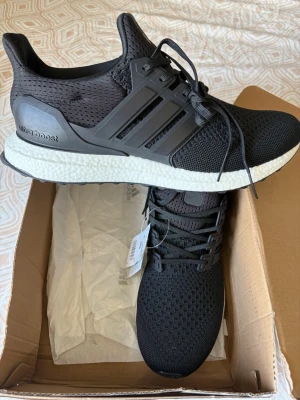 Adidas Ultraboost  - Svarta Adidas Ultra Boost sneakers med stickad ovandel och klassiska tre ränder på sidan. Skorna har en vit Boost-mellansula för extra komfort och svart yttersula. Snygg sportig design med snörning och ventilerande meshmaterial.