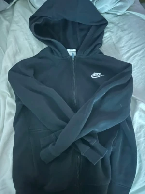 Svart Nike zip hoodie med huva - Svart hoodie från Nike med dragkedja framtill och klassisk vit logga på bröstet. Tröjan har huva och två fickor framtill. Perfekt för en chill och sportig look. Materialet är mjukt och skönt, troligtvis bomullsblandning.