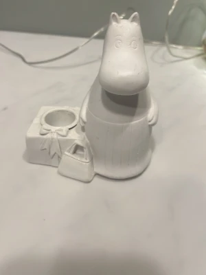Vit Mumin ljushållare i keramik - Söt ljushållare i form av Mumin, gjord i vit keramik. Figurerad med present och väska vid sidan, perfekt som dekoration eller för värmeljus. Passar dig som gillar Mumin och vill ha något unikt på skrivbordet eller hyllan.