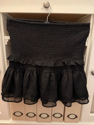 Svart volangkjol med smock - Supersöt svart kortkjol med smock upptill och dubbla volanglager nertill. Kjolen har en luftig känsla och är perfekt för dig som gillar en feminin och trendig stil. Materialet känns lätt och flowy, och volangerna ger extra rörelse.