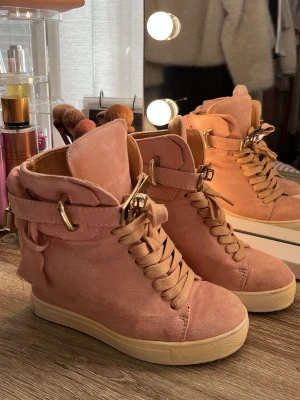 Rosa sneakers med gulddetaljer - Säljer ett par snygga rosa sneakers i mockaimitation med högt skaft och guldfärgade detaljer som spännen och lås. Skorna har breda snören och vit sula. Perfekta för dig som vill sticka ut med din stil.
