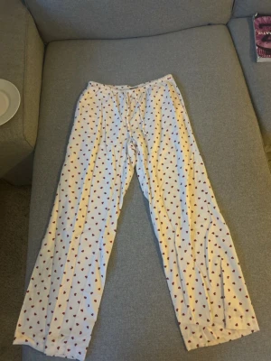 Vita pyjamasbyxor med hjärtmönster - Säljer ett par vita pyjamasbyxor med röda hjärtan över hela tyget. Byxorna har resår i midjan och är raka i modellen, perfekta för mysiga hemmakvällar. Materialet känns mjukt och bekvämt mot huden.