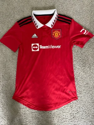 Manchester United matchtröja Adidas - Röd Manchester United matchtröja från Adidas med vit krage och svarta detaljer. Tröjan har klubbmärke på bröstet, TeamViewer-logga och klassiska Adidas-ränder på axlarna. Perfekt för fotbollsträning eller att bära på matchdag.